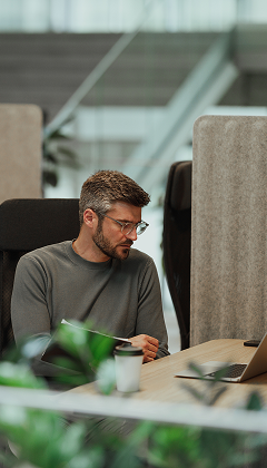 A man with glasses sits at a desk in a modern office looking at a laptop