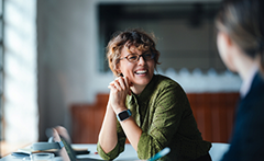A woman with curly hair and glasses smiles while sitting at a table